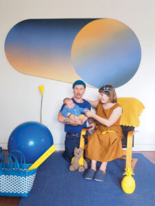 A man stands holding a baby in his arms. Next to him a woman sits, looking at the baby, with one arm around the man and one arm reaching out to the baby. The image is styled in blue and yellow, with a blue and yellow gradient graphic artwork on the wall, a yellow throw over the chair, a large blue ball on the ground, the man and the baby dressed in blue and the woman dressed in yellow. THere is a teddy bear sitting on the ground.