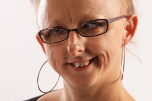 A woman with white skin, sparse short blonde hair, large hoop earrings and black rimmed glasses, smiling.
