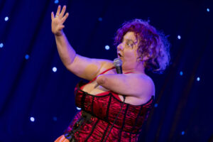 A woman is on stage, wearing a red burlesque outfit. She is holding a mic with one arm and gesturing up with the other. She has frizzy hair and a surprised look on her face.