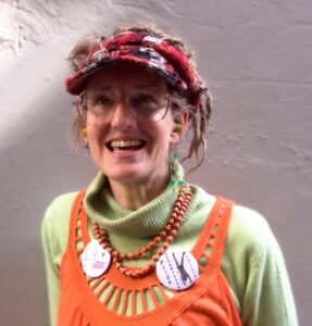 A photograph of a smiling woman facing towards the camera and shown from the chest upwards. She has white skin and light eyes, with light brown hair pulled back from her face by a red, white and black headcovering. She wears glasses and earplugs, a long sleeved and high necked light green jumper with a bright orange knitted shirt layered on top with orange beads. She has large badges on her chest. 