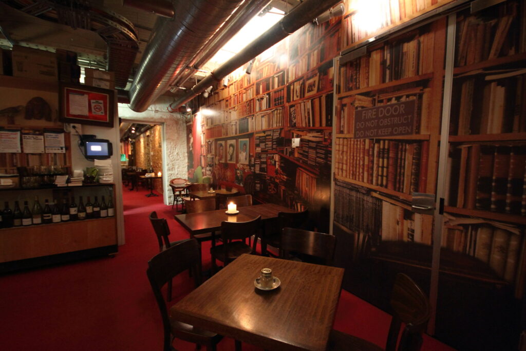 A photograph of the Moat Cafe, showing a subterranean venue with tables and chairs in front of bookshelves on all walls.