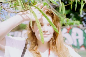 Photograph of a person partially hidden by green foliage cascading down in front of them. They have one hand raised up to their forehead and are looking to the left of the camera. The figure has pale skin, long light brown hair and a light shirt