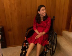 A smiling woman with long black hair sits in her wheelchair. Behind her is a wall of wood panelling. She wears a red dress.