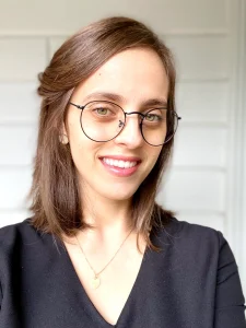 A portrait of Liel smiling at the camera. Liel is a white person with olive skin and shoulder-length brown hair. She is wearing a black top and black thin glasses.