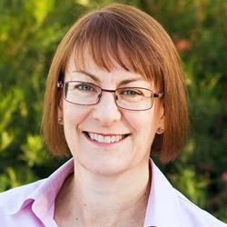 A woman with red hair looks at the camera. She wears glasses and a pink collared shirt.